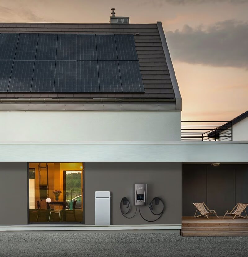 A modern house with rooftop solar panels and a dcbel home energy unit with EV charging cables mounted on the exterior wall
