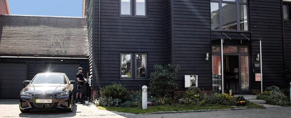 An electrician working at an EV charge point on the driveway of a dark timber-clad house with a black electric car parked outside