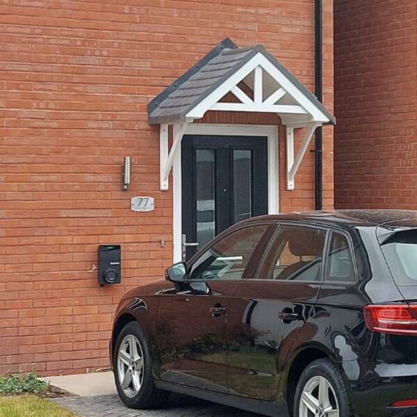 A black wall-mounted EV charge point installed on the brick exterior of a house beside a parked black car