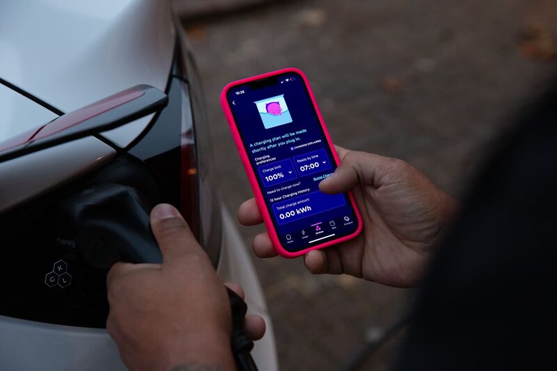 A person plugging a charging cable into an electric car while holding a phone showing an EV charging app