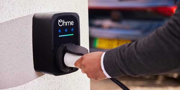 A person plugging a charging cable into a black Ohme wall-mounted electric car charger on a white wall