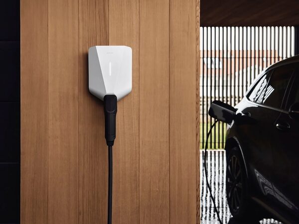 A white Easee wall-mounted EV charger with cable on a wooden wall beside a car charging in a carport