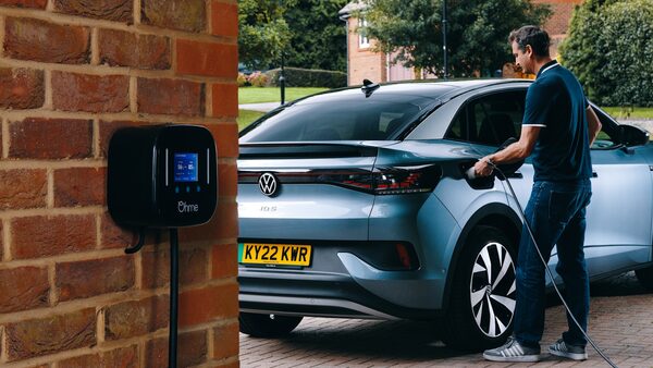 A man plugging a charging cable into a blue Volkswagen ID.5 electric car on a driveway next to a black Ohme wall-mounted EV charger on a brick wall