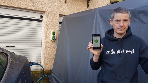 A man holding a smartphone showing a charging app next to a wall-mounted EV charger plugged into a car outside a garage