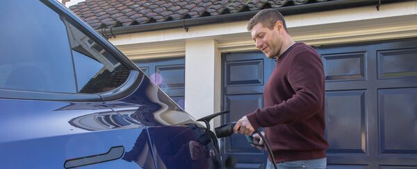 A man plugging a charging cable into a dark blue electric car on a driveway outside a house with a garage