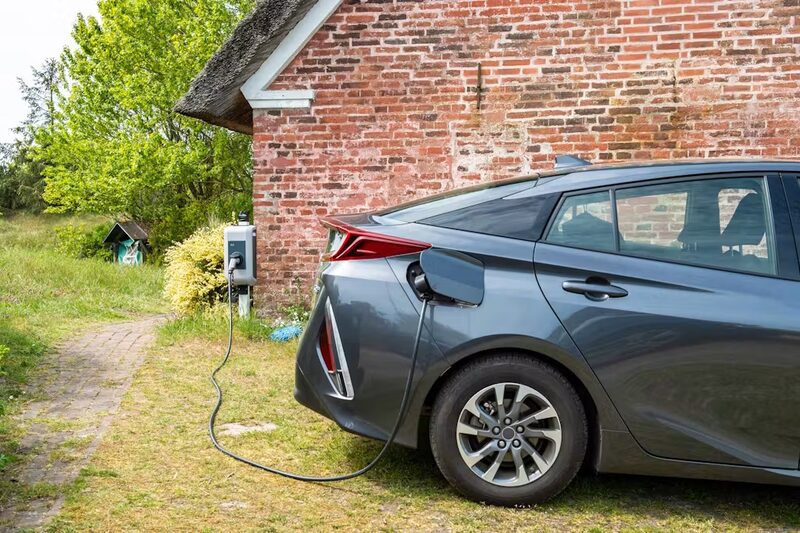 A grey Toyota Prius plug-in hybrid charging from a wall-mounted charger outside a brick house with a thatched roof