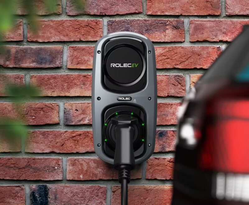 A Rolec EV wall-mounted electric car charge point with a plugged-in cable on a brick wall beside a car