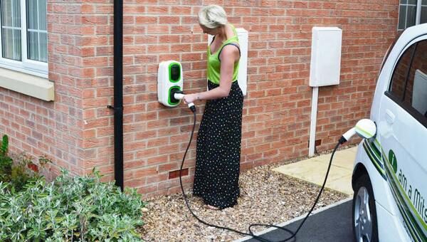 A woman plugging a charging cable into a green and white Rolec Homecharge WallPod EV charger on a brick house wall, with a white electric car parked on the driveway
