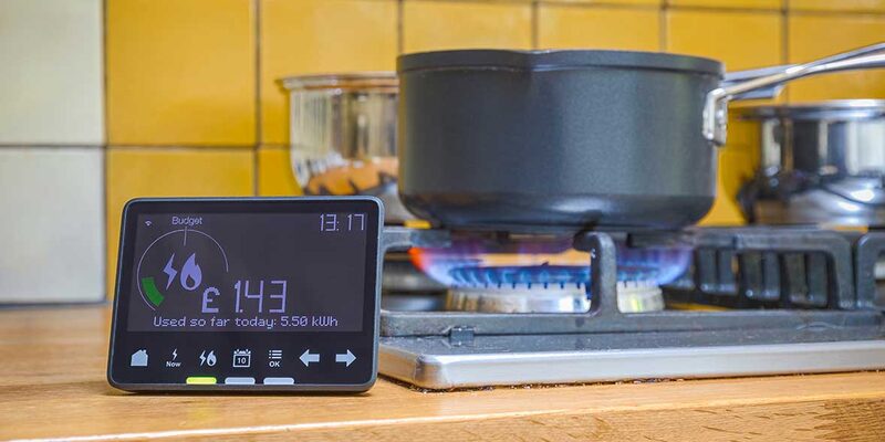 A smart meter in-home display showing energy use and cost sitting on a kitchen worktop beside a lit gas hob with a saucepan