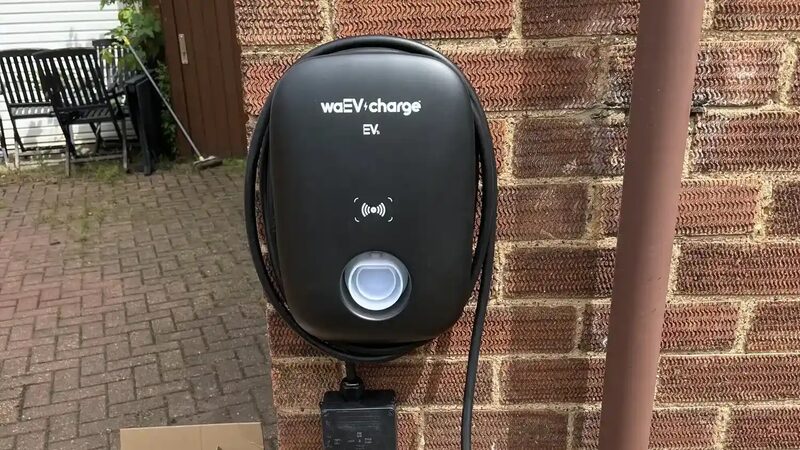 A black waEV-charge EV home charger mounted on a brick house wall with coiled cable