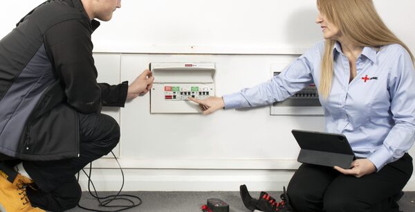Two electricians inspecting a wall-mounted consumer unit fuse board, one holding a tablet