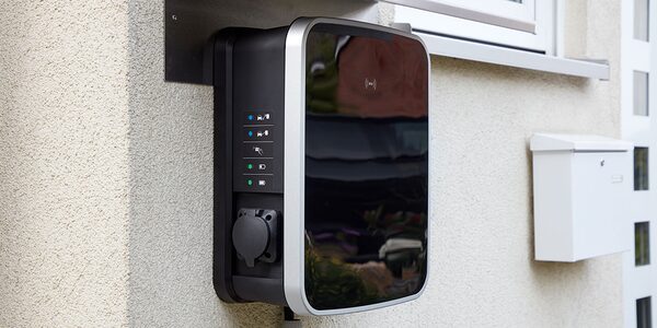 A black and silver wall-mounted electric car chargepoint with status lights and a socket cover, fitted to the exterior wall of a house next to a white postbox