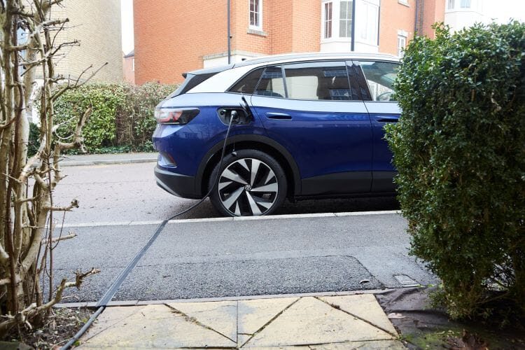 A blue Volkswagen ID.4 electric car parked at the kerb with a charging cable running across the pavement to the charge port