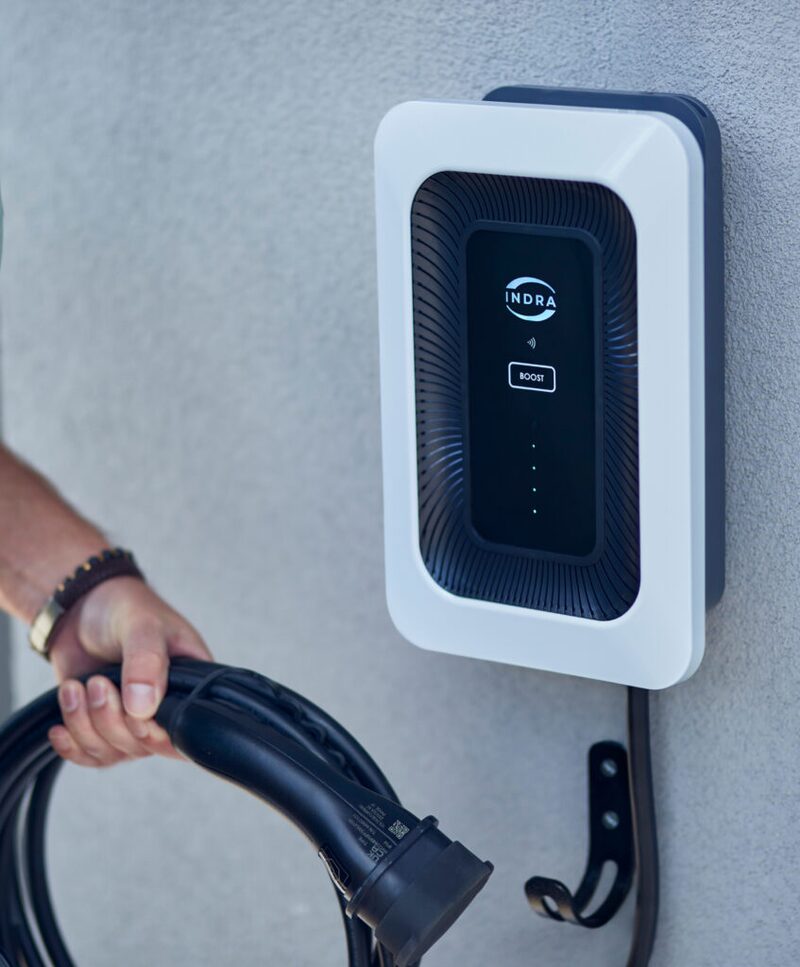 A person holding an EV charging cable next to a white Indra Smart Pro wall-mounted electric car charger on an exterior wall