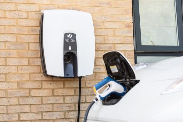 A white Indra wall-mounted EV charger on a brick wall with a charging cable plugged into a white car