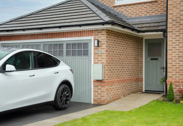 A white electric car parked on a driveway beside a brick house with an Andersen EV wall charger mounted next to the garage