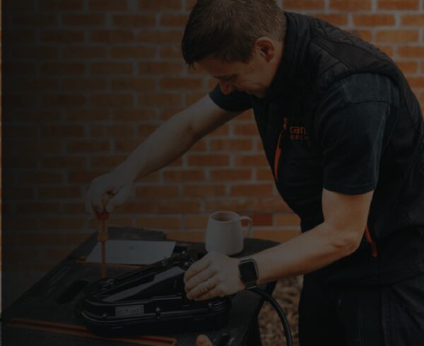 An electrician in a dark work vest using a screwdriver on an EV charger unit outdoors against a brick wall