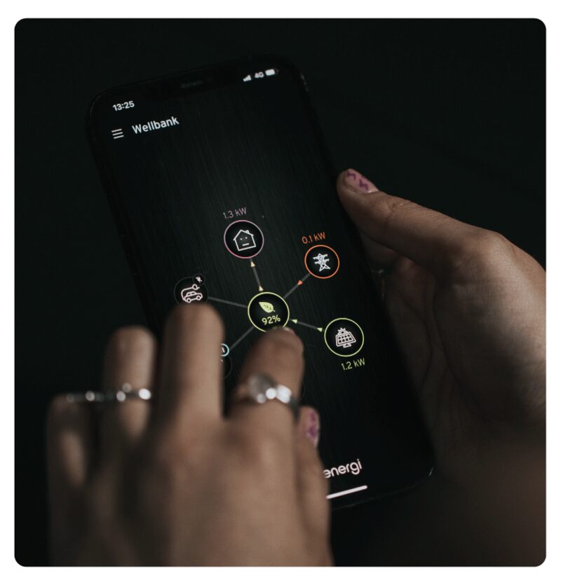A person's hands holding a smartphone showing the myenergi app with an energy flow diagram linking home, grid, EV and solar icons