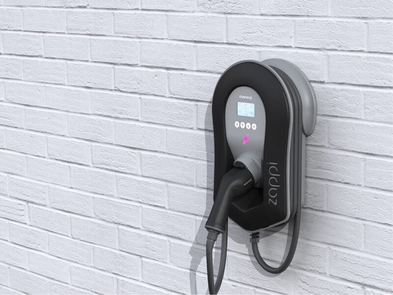 A myenergi zappi EV charge point mounted on a white brick wall with its charging cable holstered