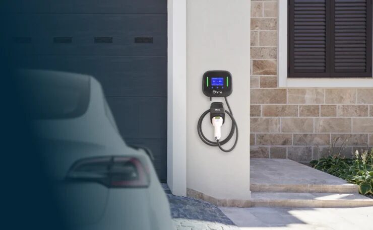 An Ohme wall-mounted EV charger with coiled cable installed on a house wall beside a garage, with a car in the foreground