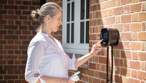A woman touching the screen of an Ohme Home Pro EV charger mounted on a brick house wall while holding the charging cable