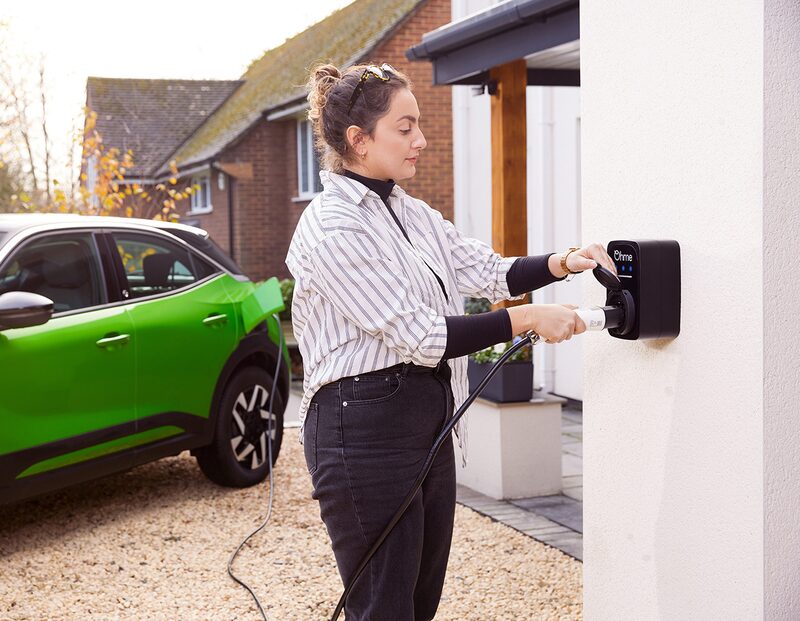 A woman plugging a charging cable into a black Ohme wall-mounted EV charger outside a house, with a green electric car parked on the driveway
