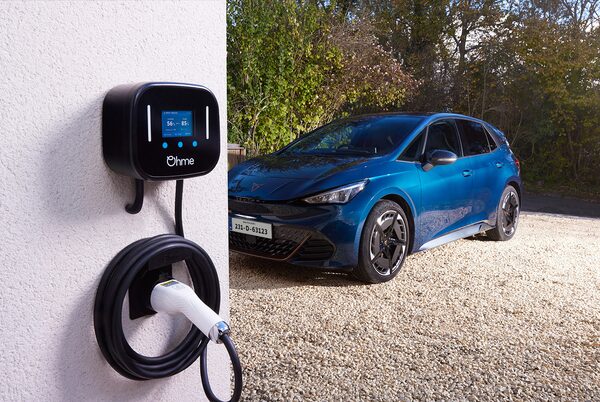 An Ohme wall-mounted EV charger with coiled cable on a house wall next to a blue Cupra electric car on a gravel driveway