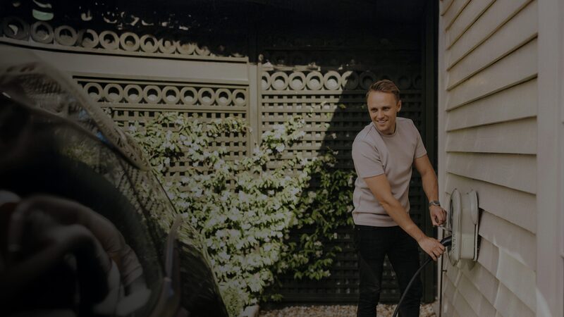 A man plugging a charging cable into a wall-mounted EV charger outside a house next to a car