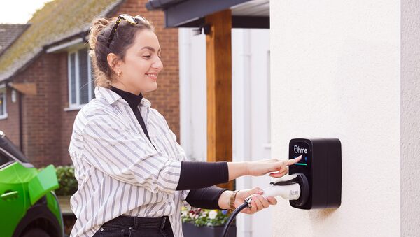 A woman plugging a charging cable into a black Ohme wall-mounted EV charger outside a house