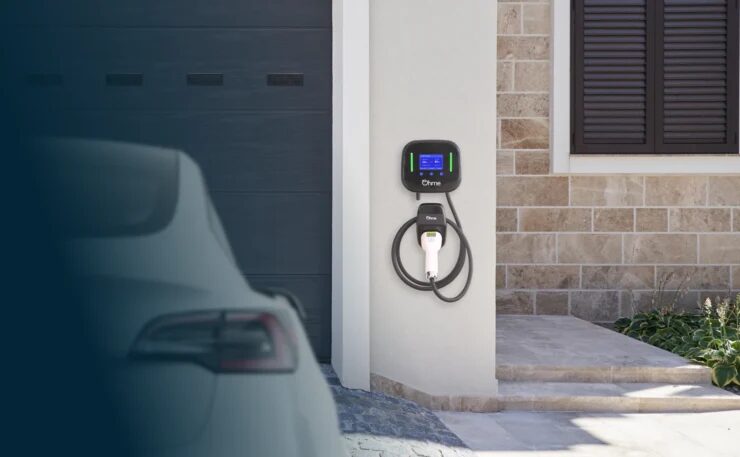 An Ohme wall-mounted home EV charger with tethered cable installed on a house wall beside a garage, with a car parked in the foreground