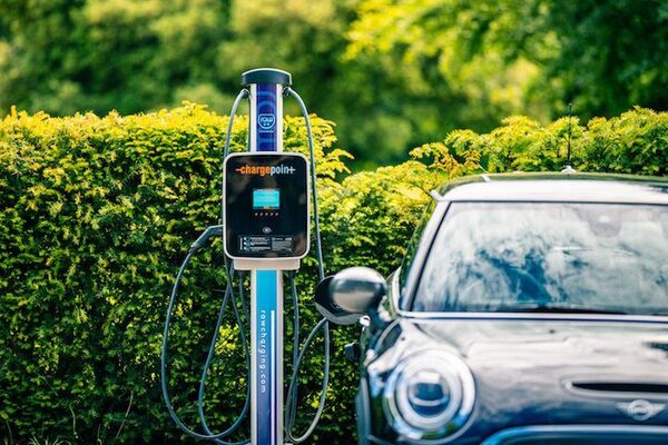 A ChargePoint public EV charging post with cables beside a parked car in front of a green hedge