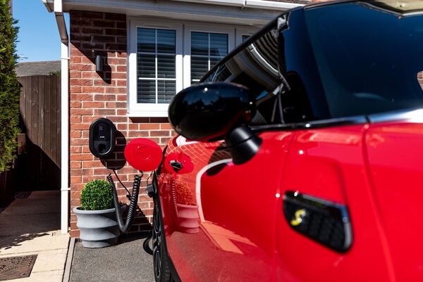 A red Mini Electric plugged into a wall-mounted EV charger on a brick house driveway