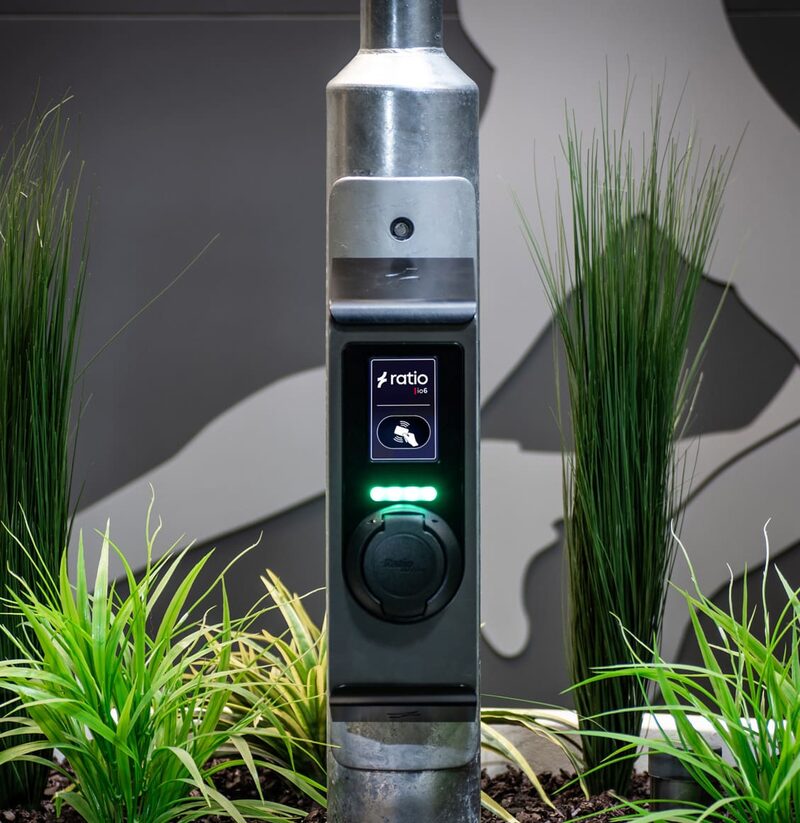A Ratio io6 street EV charger mounted on a metal post with a lit socket and display, surrounded by ornamental grasses