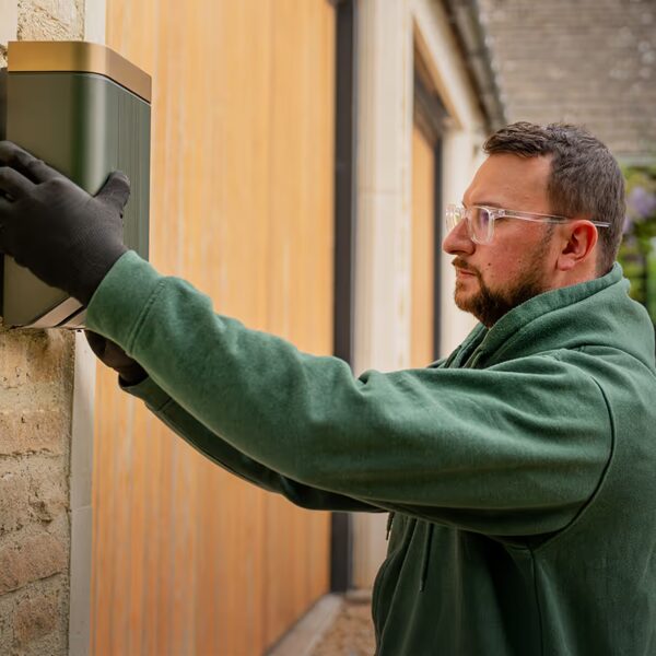 A man in a green hoodie and black gloves installing a green wall-mounted EV charge point on a brick house wall
