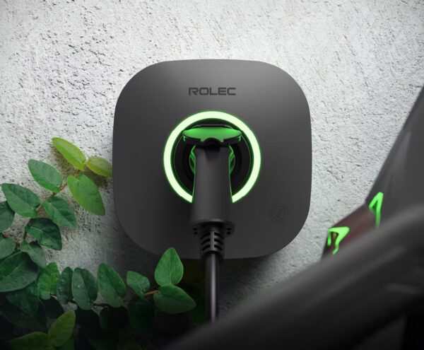 A Rolec wall-mounted EV charge point with a glowing green ring and a charging cable plugged in, mounted on a white wall beside a plant