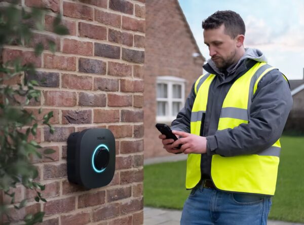 An installer in a hi-vis vest using a phone next to a black Rolec EVO EV charge point mounted on a brick house wall