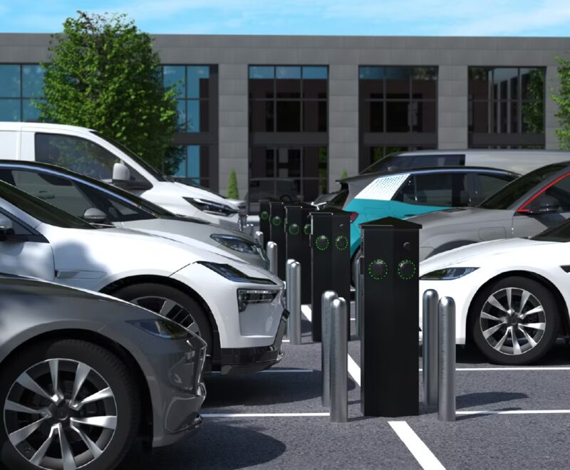 A row of black Rolec HEX electric car charge posts in a car park with parked cars and an office building behind