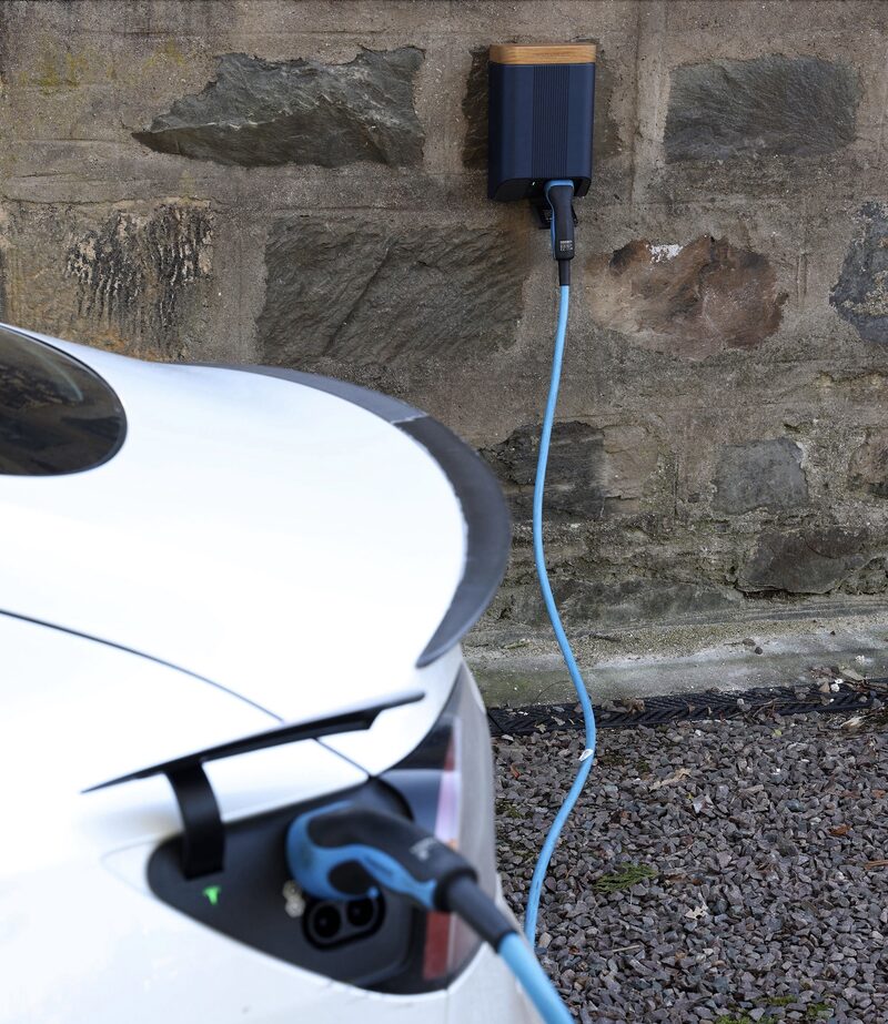 A wall-mounted Simpson &amp; Partners EV charger with a blue cable charging a white car outside a stone building