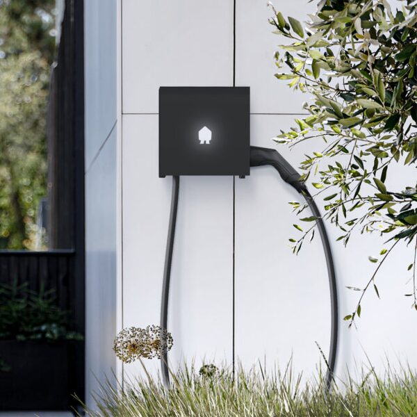 A black Smappee wall-mounted EV charger with cable and holster installed on a white exterior wall beside greenery