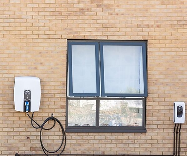 A white Indra EV charge point with coiled cable mounted on a brick house wall beside a window, with another small white unit to the right