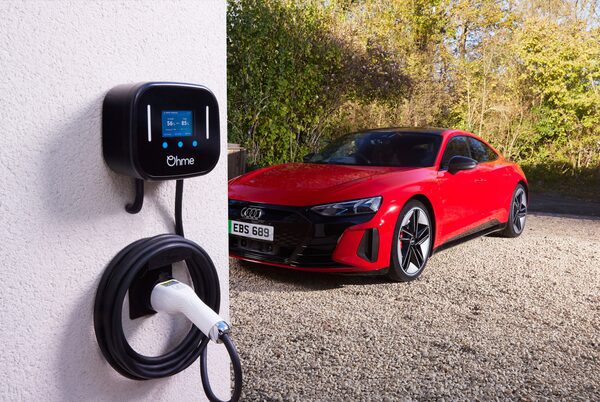 A black Ohme tethered EV wall charger mounted on a white wall with coiled cable, next to a red Audi e-tron GT parked on a gravel driveway