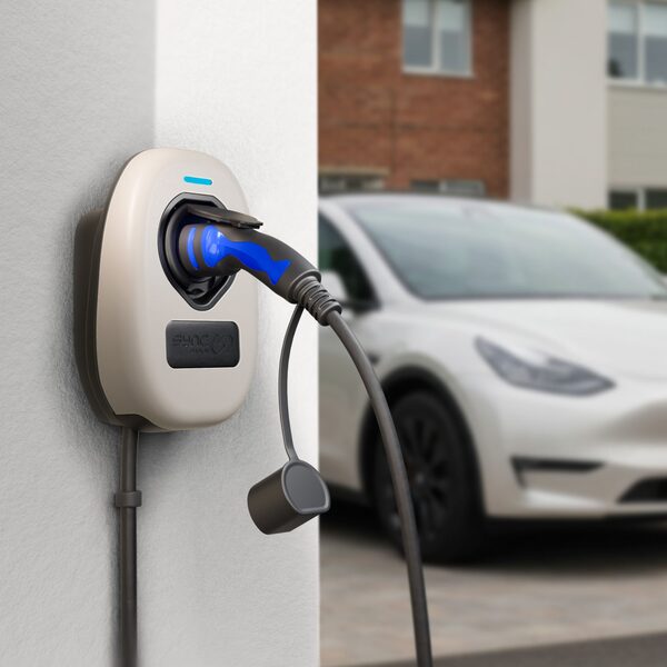 A Sync Energy wall-mounted EV charger with a tethered cable plugged in, on a house wall with a white electric car parked in the driveway behind