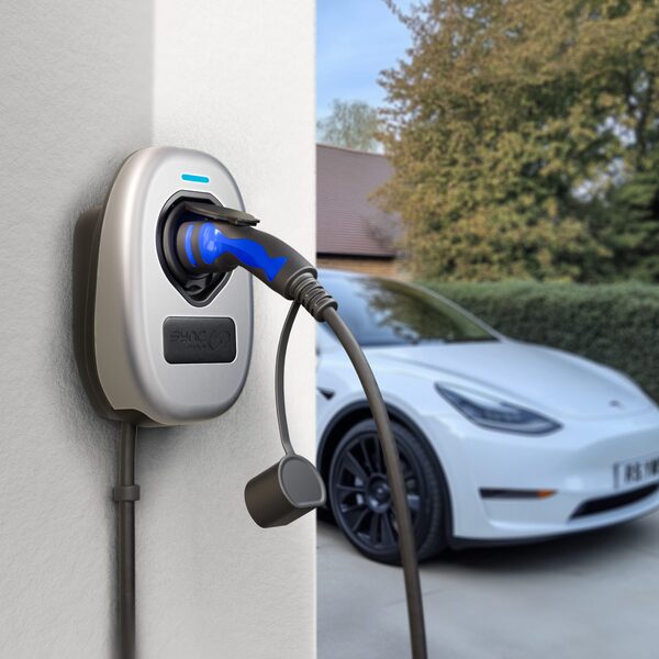 A metallic silver Sync Energy wall-mounted EV charger with a blue charging cable plugged in, on a house wall beside a white electric car on a driveway