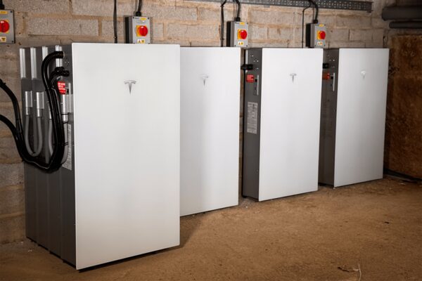 Four Tesla Powerwall home battery units installed in a row against a brick wall in a garage or cellar