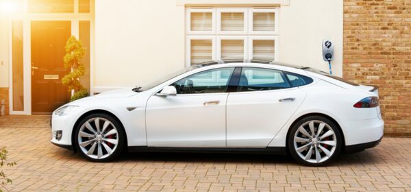 A white Tesla Model S parked on a driveway outside a house, plugged into a wall-mounted EV charge point on the brick wall