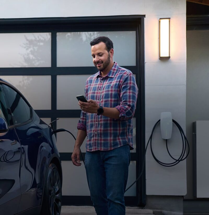 A man checking his phone next to an electric car charging from a Tesla Wall Connector mounted on a garage wall