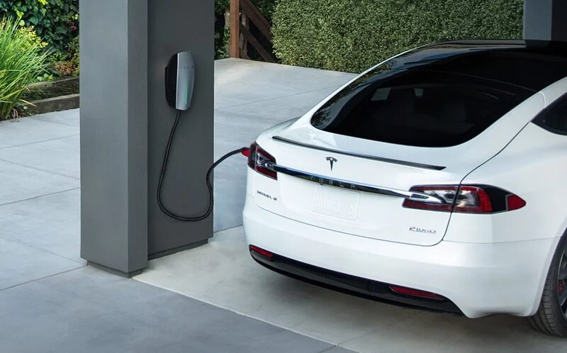 A white Tesla Model S plugged into a Tesla Wall Connector mounted on a grey pillar outside a home