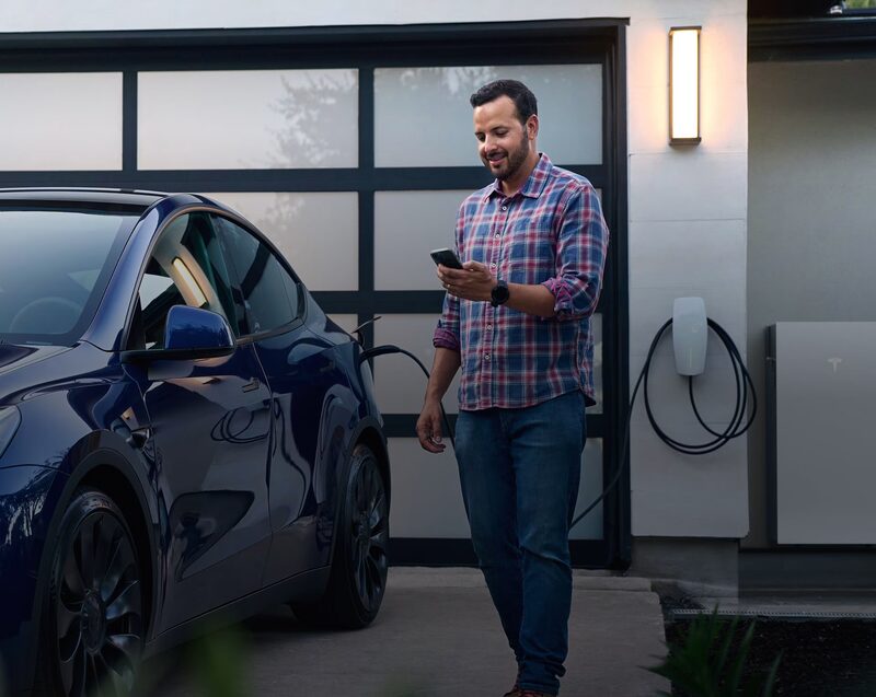 A man checking his phone beside a blue Tesla plugged into a wall-mounted Tesla Wall Connector EV charger outside a garage