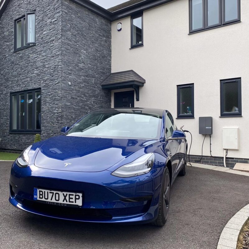 A blue Tesla Model 3 charging from a wall-mounted EV charger outside a modern UK house