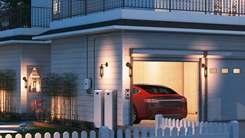 A Tesla Powerwall battery and EV charger mounted on the outside wall of a modern house at dusk, with a red car parked in the garage